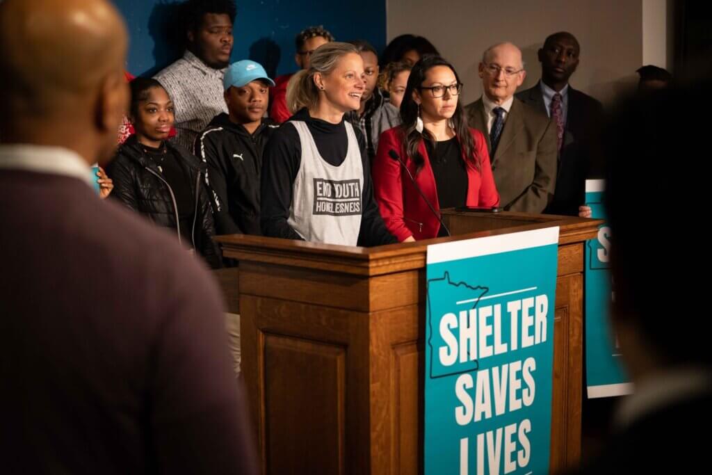 CEO Beth Holger behind podium with sign that reads "Shelter Saves Lives"